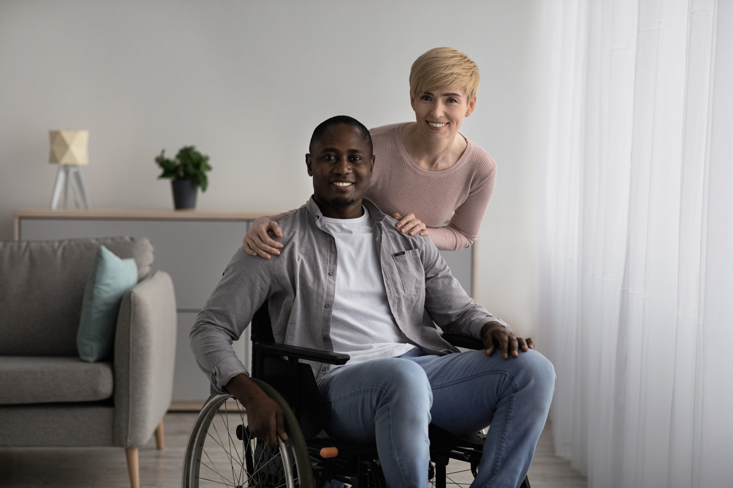 Rehabilitation clinic and patient care, home nurse or multiracial family. Mature caucasian lady in casual and Happy adult african american husband in wheelchair, in living room or clinic interior